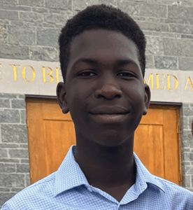 Student has short dark hair, is wearing a light blue shirt and is standing in front of a stone building.