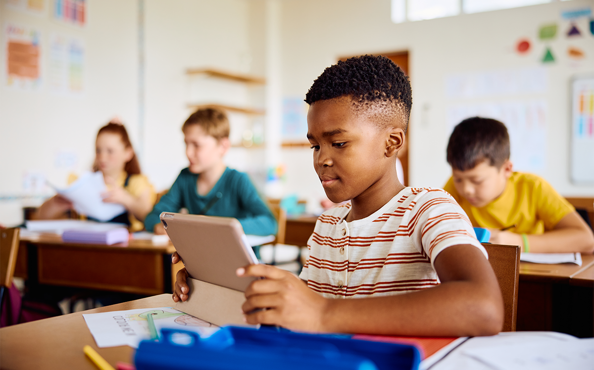 Photo of young students working at their desks in a classroom, with one student looking at a tablet in the foreground.