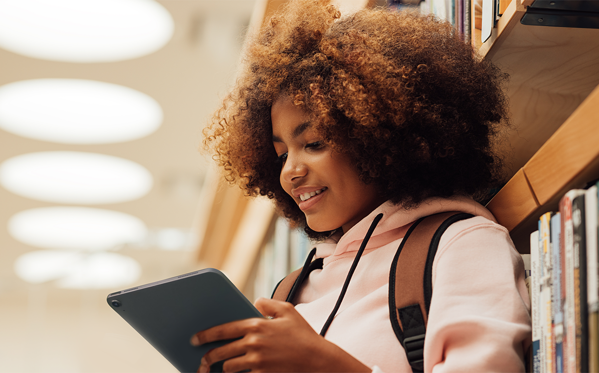 Photo of a student standing in front of stacks of books, wearing a backpack and engaging enthusiastically with a tablet.