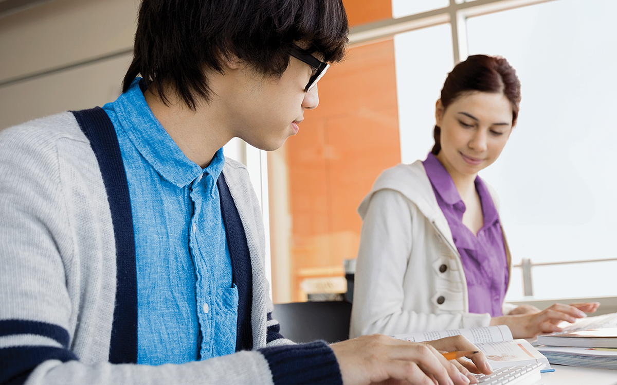 Photo de deux élèves adolescents assis devant une fenêtre, tapant sur des claviers blancs, des cahiers à côté d’eux.