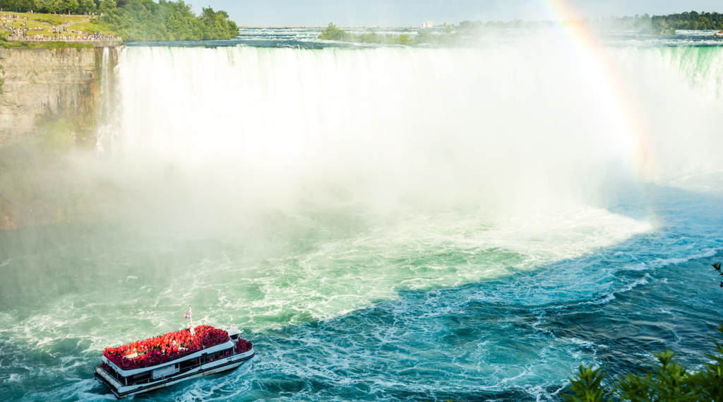 Red and white tour boat approaching Niagara Falls amid heavy mist and a rainbow on a sunny day