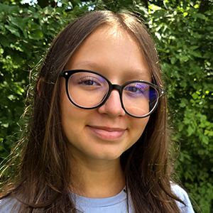 Élève portant des lunettes et un t-shirt bleu pâle, souriant légèrement à la caméra, à l’extérieur devant des arbres feuillus verts.