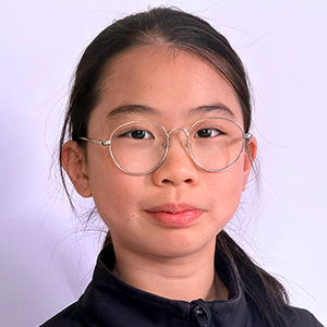 Élève aux longs cheveux foncés attachés, portant des lunettes rondes et un veston noir à fermeture éclair devant un fond pâle uni.