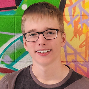 Élève portant des lunettes et un t-shirt marron, devant un mur de graffitis verts, orange et jaunes.