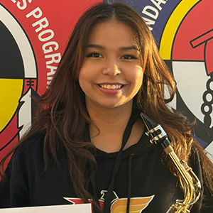 Élève tenant un saxophone et une enveloppe, devant deux bannières colorées aux motifs circulaires rouges, jaunes, noirs et blancs.