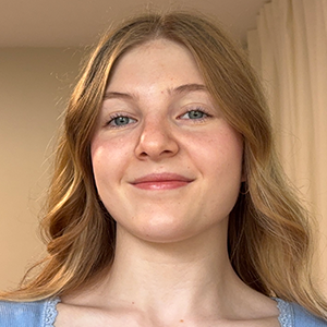 Élève aux cheveux brun clair portant un haut bleu pâle, souriant légèrement à la caméra, devant un mur beige et des rideaux crème.