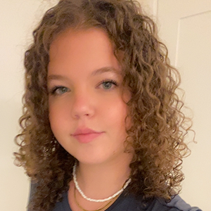 Élève aux cheveux bruns bouclés portant un haut noir et des colliers superposés, souriant légèrement devant un mur pâle et une porte.
