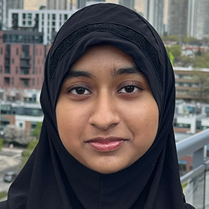 Élève portant un hijab noir et un haut beige sur un balcon, avec des immeubles et un ciel nuageux en arrière-plan.