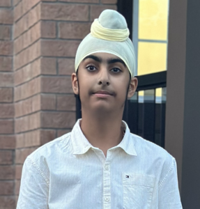 Student with a neutral expression, wearing a light-coloured turban and a white button-up shirt, standing outdoors against a brick wall and window.