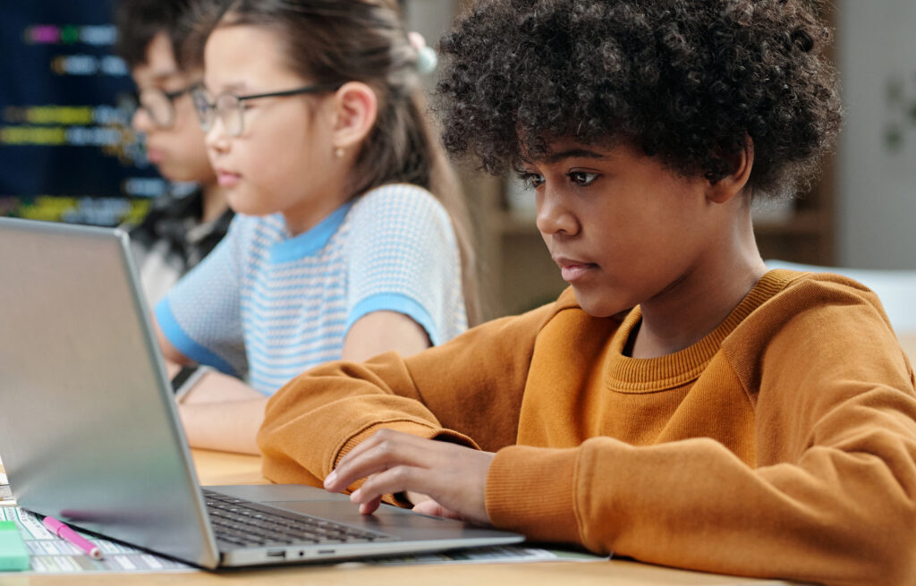 Photo of young students working on laptops in a classroom, one student in the foreground typing on a laptop.