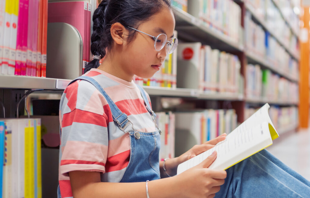 Photo of a young student sitting by bookshelves in a library, reading a book.