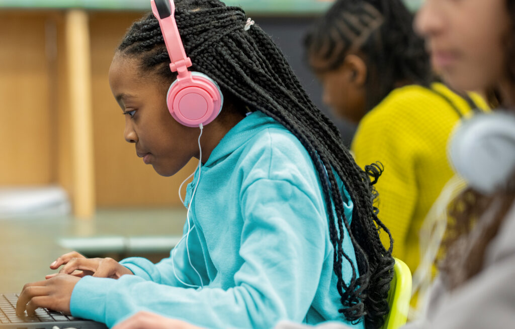 Photo of a young student wearing headphones and typing on a laptop in a classroom, other students in the background.