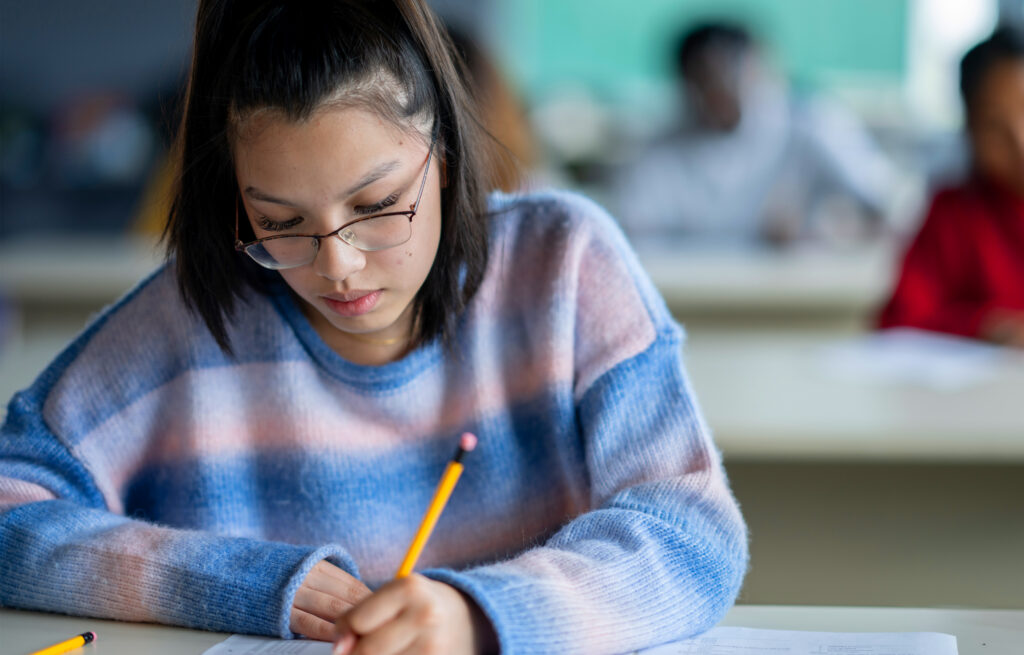 Photo of an adolescent student wearing glasses and writing with a pencil at a desk, other students in the background.