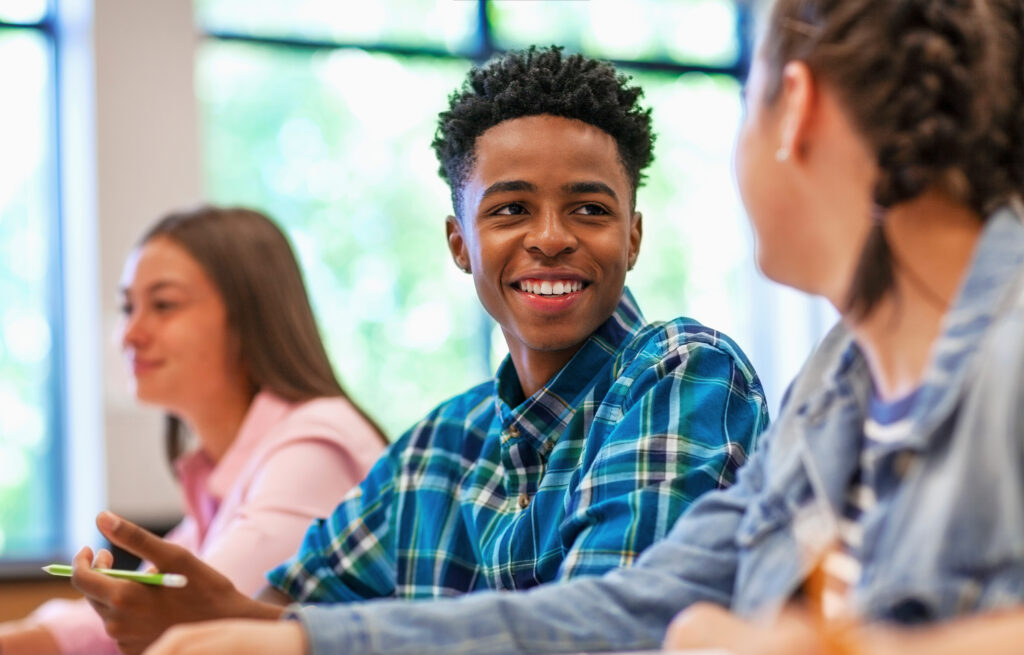 Photo of adolescent students sitting together in a classroom, one student smiling and engaging with another student.