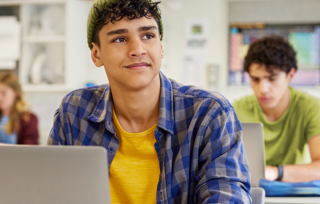 Photo of adolescent students sitting at desks with laptops in a classroom, one student in the foreground looking up.