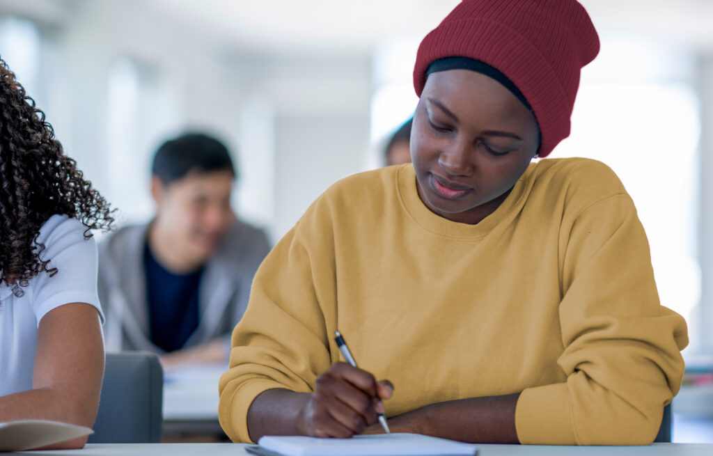 Photo of adolescent students sitting at desks, one student in the foreground writing with a pen in a notebook.